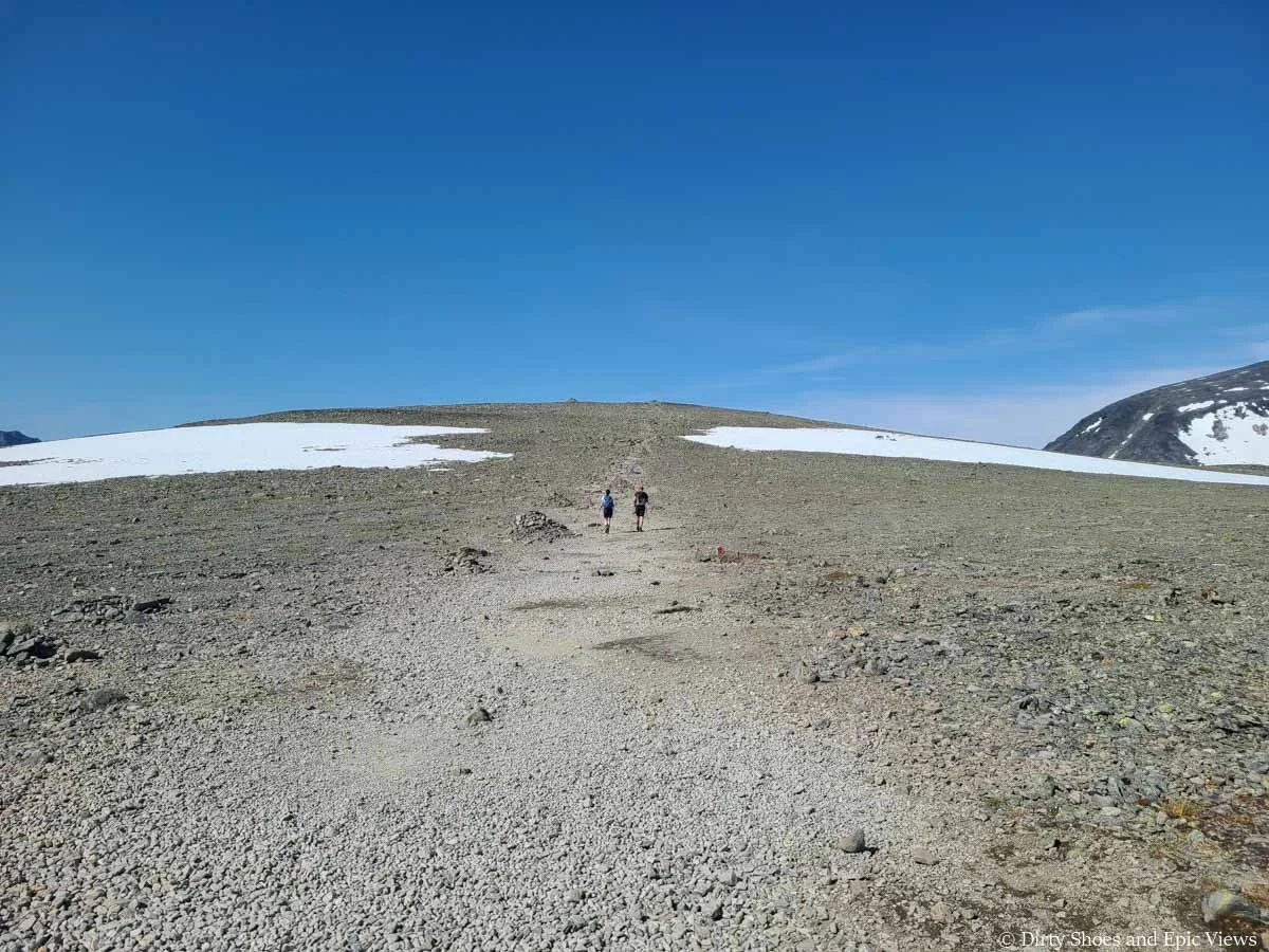 A pair of hikers walks a faint herd path through a rocky landscape with snow patches on the Besseggen Ridge trail in Norway