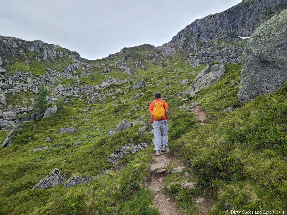 A hiker ascends a narrow dirt path through a grassy meadow towards a rocky headwall on the Reinanuten trail in Norway