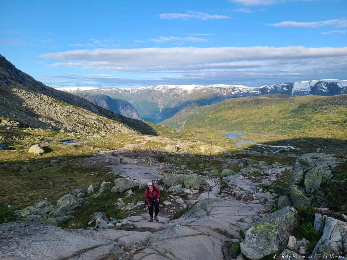 A hiker ascends a rocky path above a lake-spotted meadow with snowcapped mountains in the distance on the Trolltunga hike