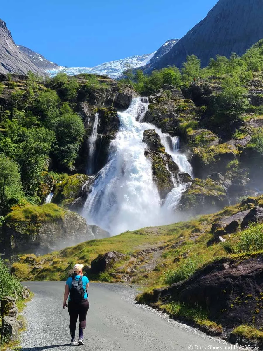 A hiker walks a path towards a powerful waterfall along the Briksdal Glacier trail in Norway