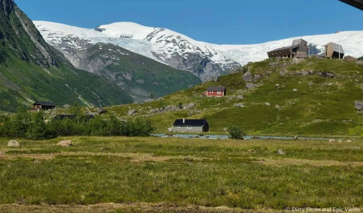 Some small buildings sit on top of a grassy hill overlooking an ice cap at the Austerdalsbreen trailhead in Norway