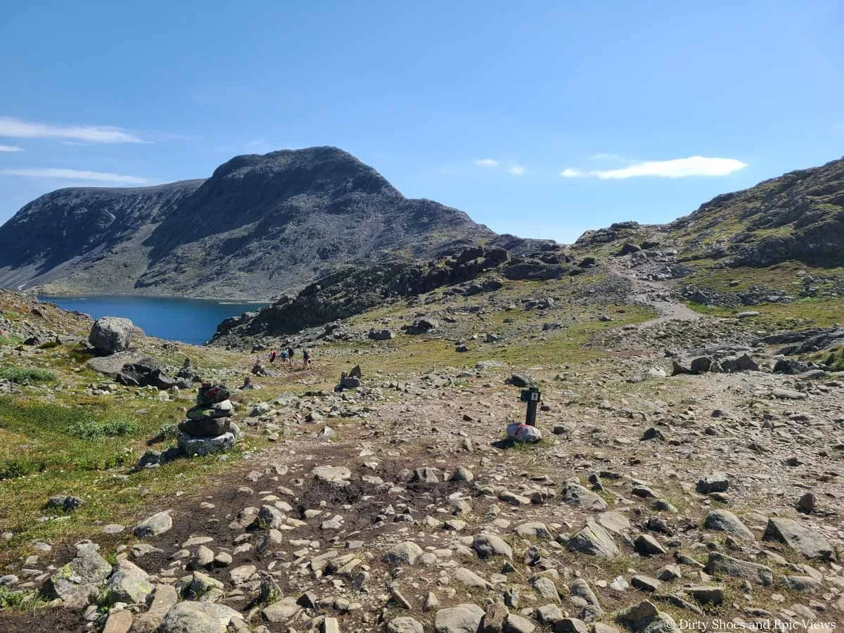 A wide rocky trail leads down through a grassy meadow towards a lake on the Besseggen Ridge trail in Norway