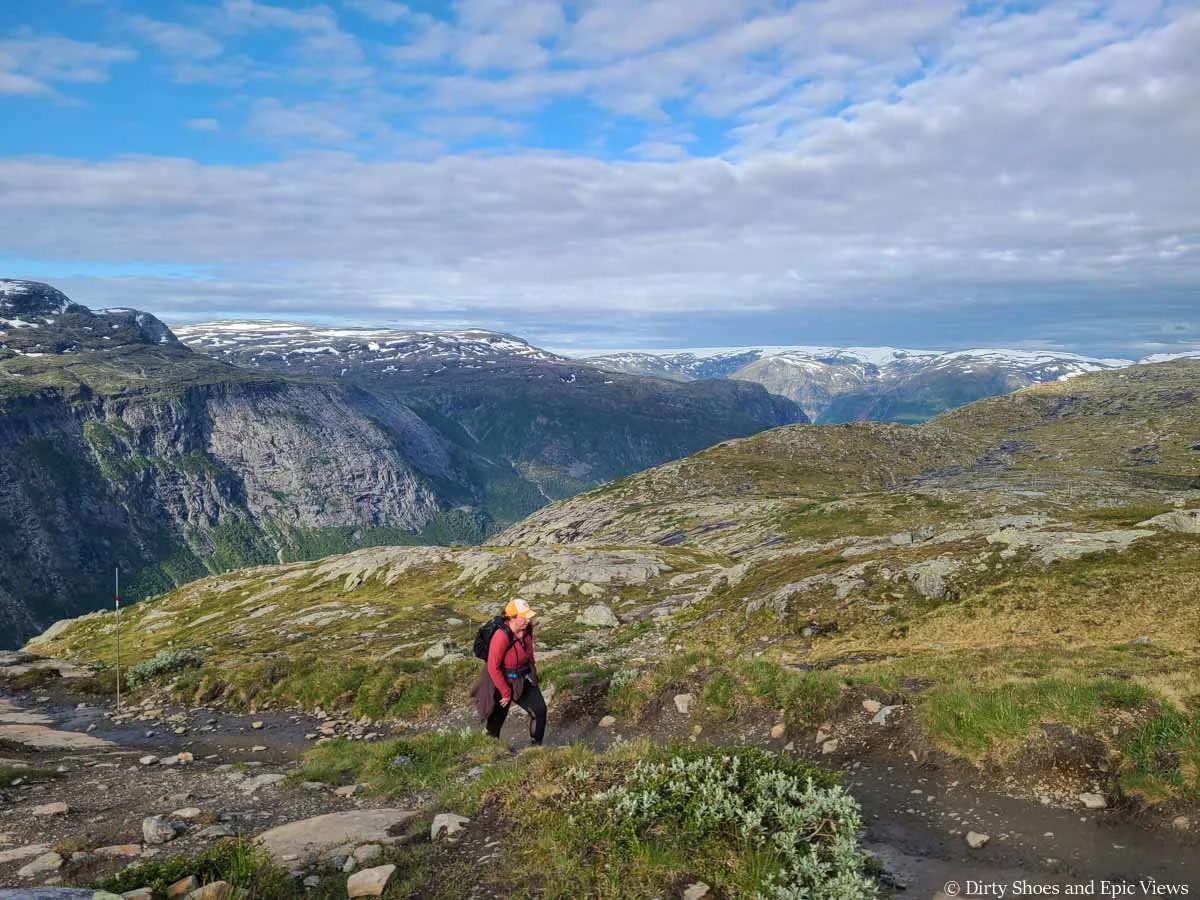 A hiker walks along a dirt path through a grassy meadow overlooking snow covered mountains on the Trolltunga hike