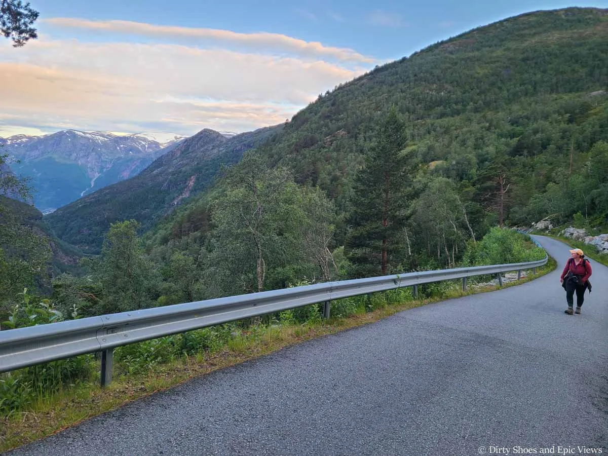 A hiker ascends a paved road and looks out to distant snowcapped mountains on the way to the Trolltunga trailhead