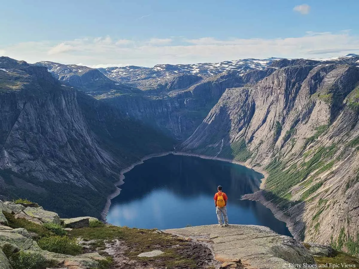 A hiker stands on a rocky viewpoint overlooking a blue lake surrounded by steep cliffs and snow covered mountain tops on the Trolltunga trail
