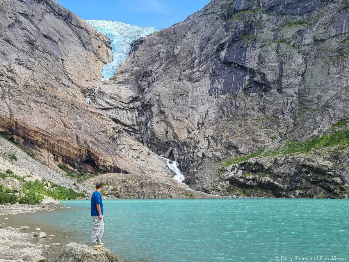 A hiker stands on a rock on the shore of a blue lake beneath a glacier at Briksdalsbreen in Norway