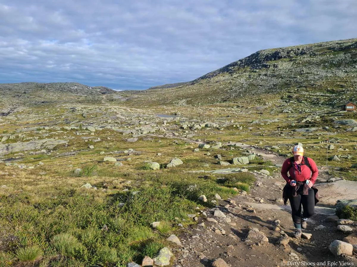 A hiker walks along a dirt path through a rocky meadow on the Trolltunga hike