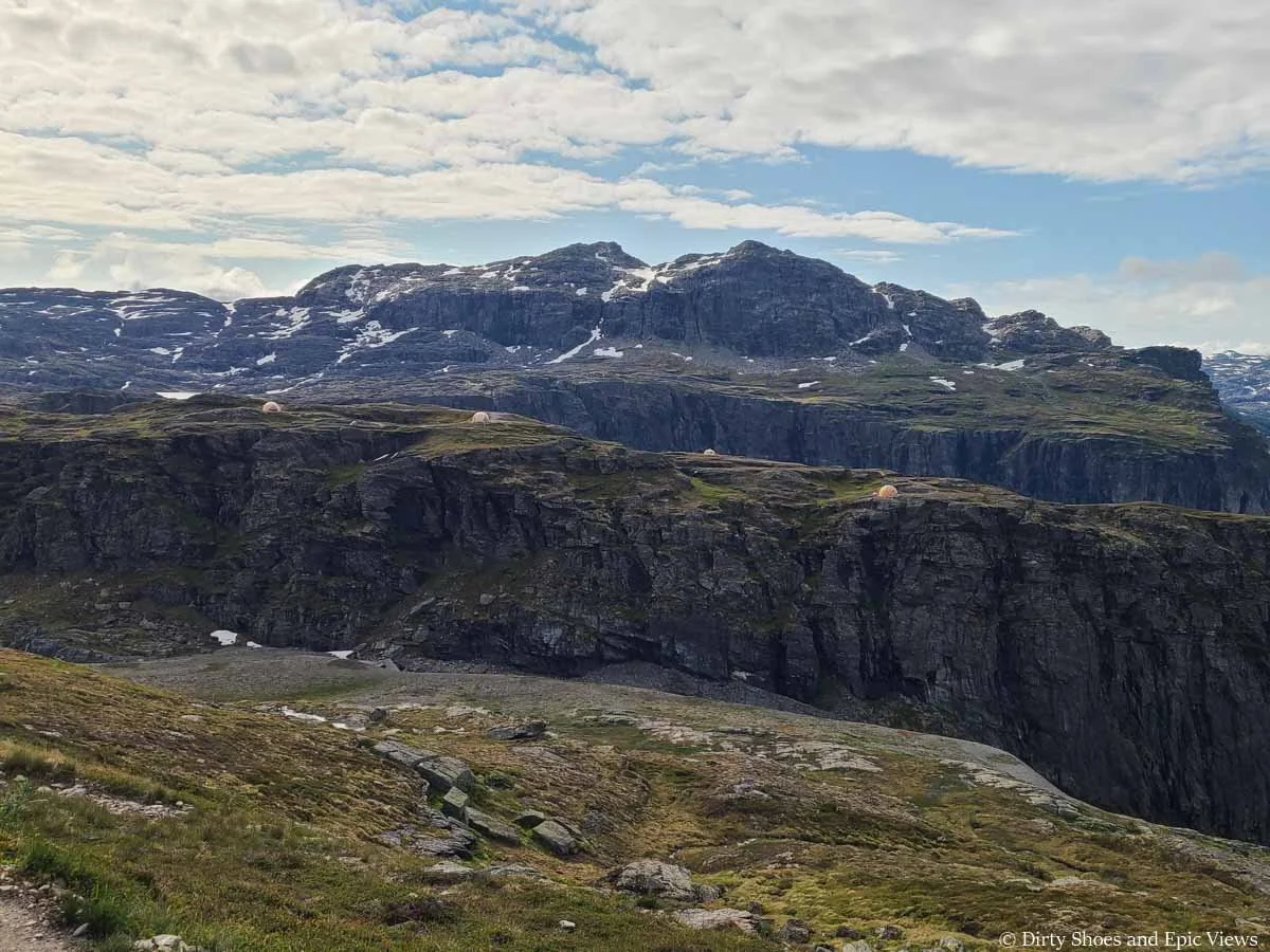 A rocky mountain sits above cliffs and meadows dotted with tents along the Trolltunga trail