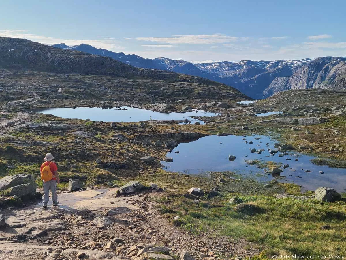A hiker descends a rocky path towards still tarns in a rocky meadow on the Trolltunga hike