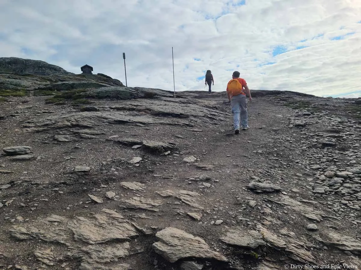 A pair of hikers ascends a steep path up a rocky slope towards a shelter along the Trolltunga hike