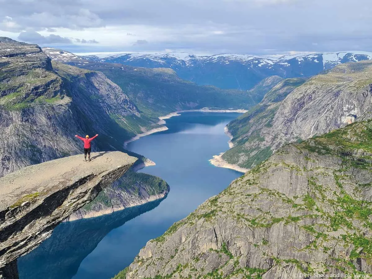 A hiker stands on a rocky outcropping overlooking a blue lake and mountains at Trolltunga in Norway