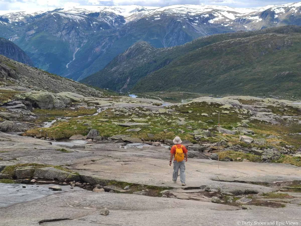 A hiker descends a granite slab towards snowcapped mountain views on the Trolltunga trail