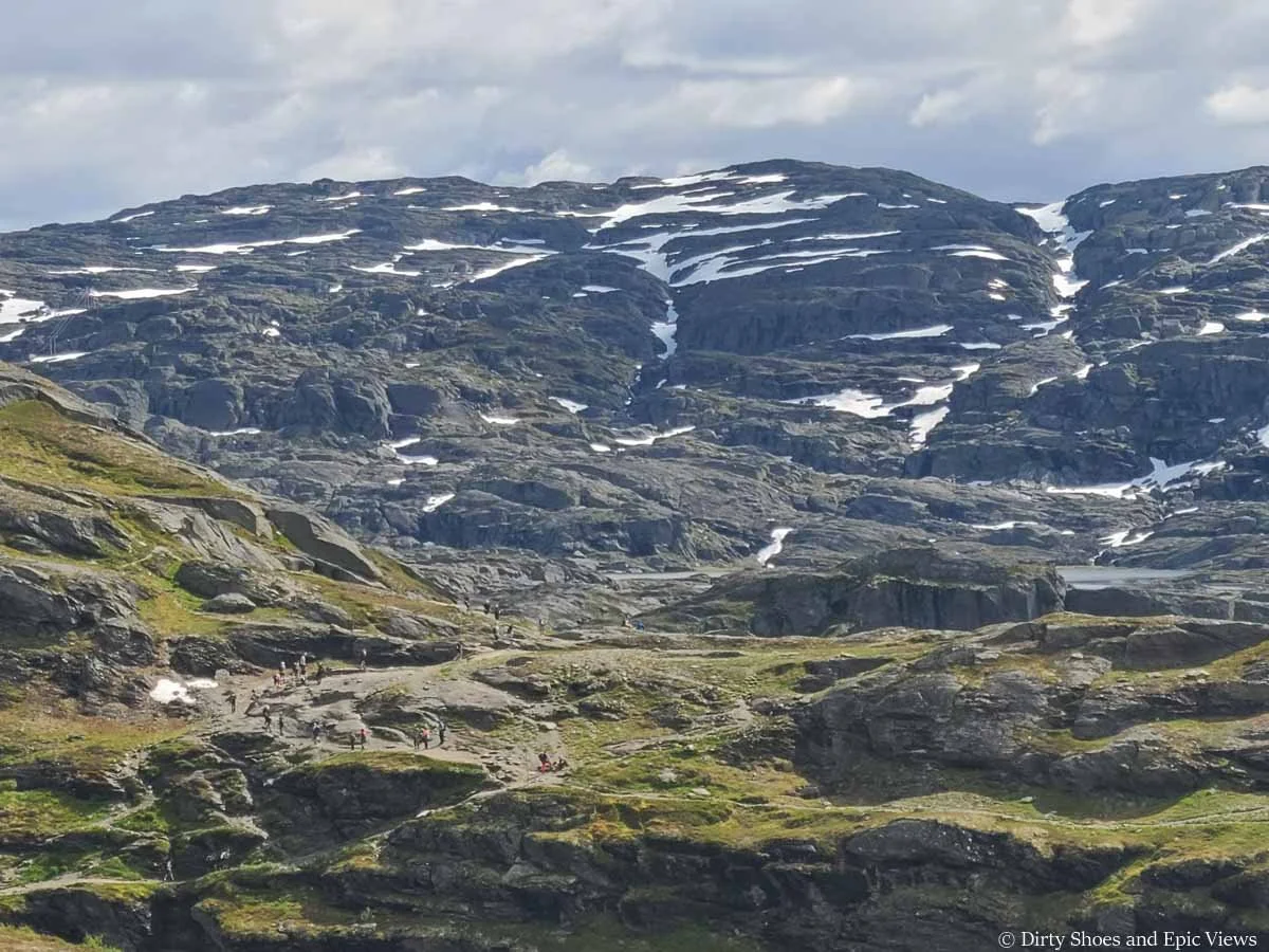 A line of hikers can be seen on a dirt trail in front of massive mountain views along the Trolltunga hike