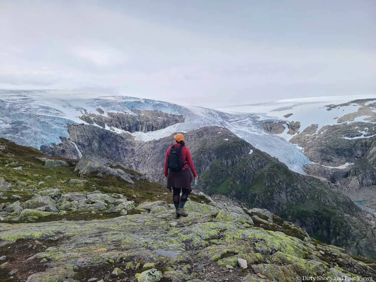 A hiker walks in front of views of mountains topped with a large icecap as seen from the Reinanuten hike in Norway