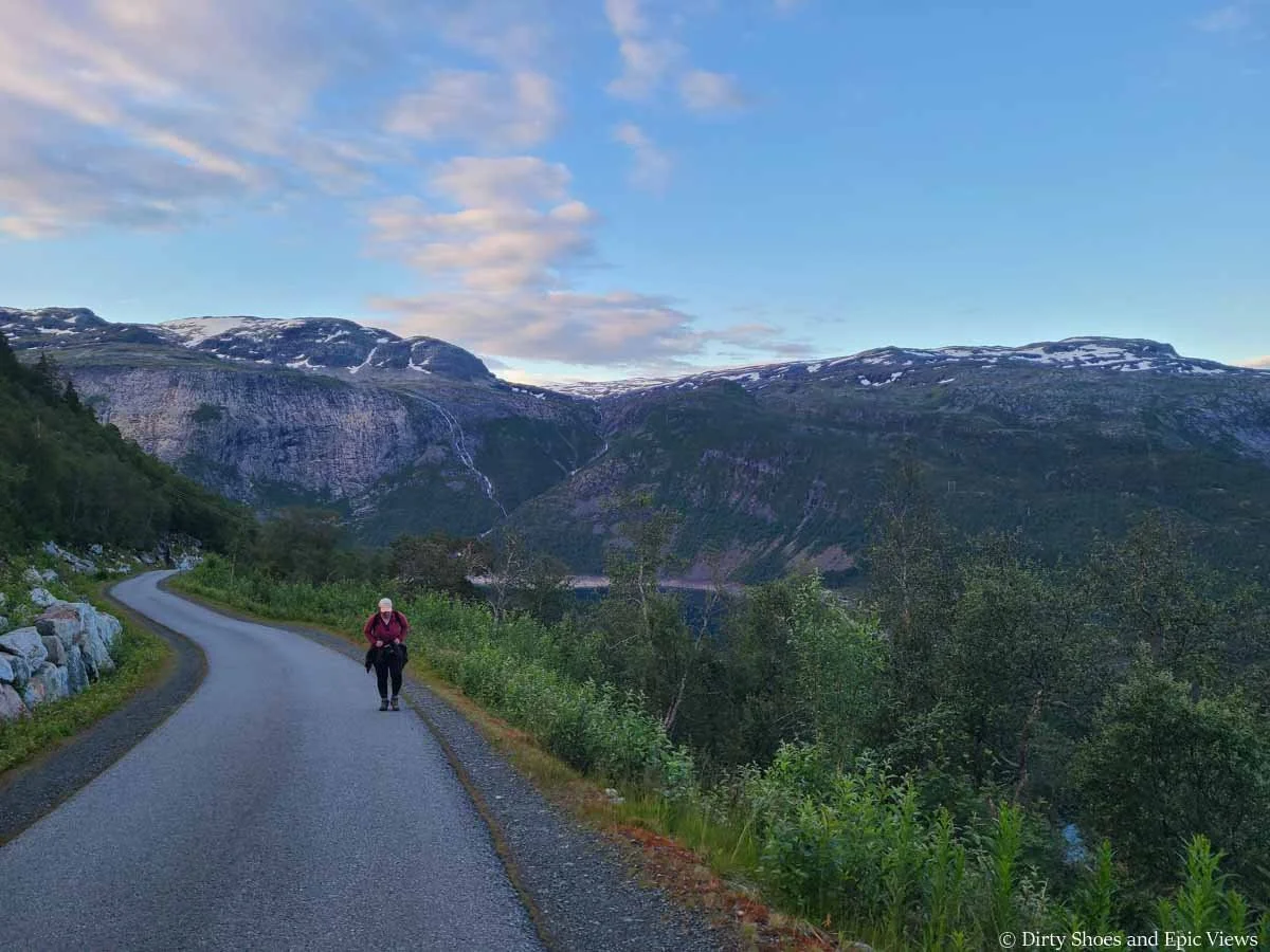 A hiker ascends a narrow paved road in front of mountain views on the way to the Trolltunga trailhead in Norway