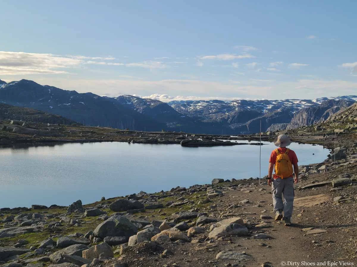 A hiker walks towards a large still lake with a mountain backdrop on the Trolltunga hike
