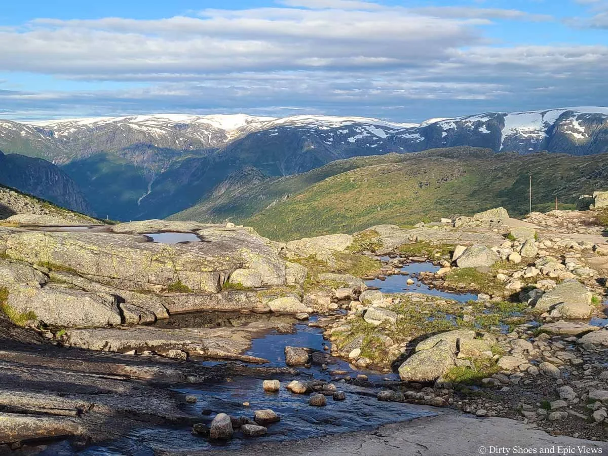 Small tarns spot a rocky landscape with distant snowcapped mountains as seen from the Trolltunga hike