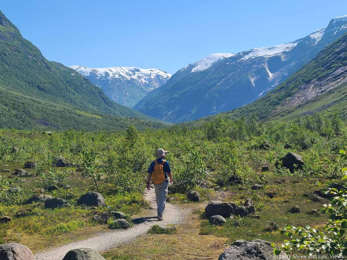 A hiker walks a narrow dirt path through a grassy meadow towards snow capped mountains on the Austerdalsbreen hike in Norway