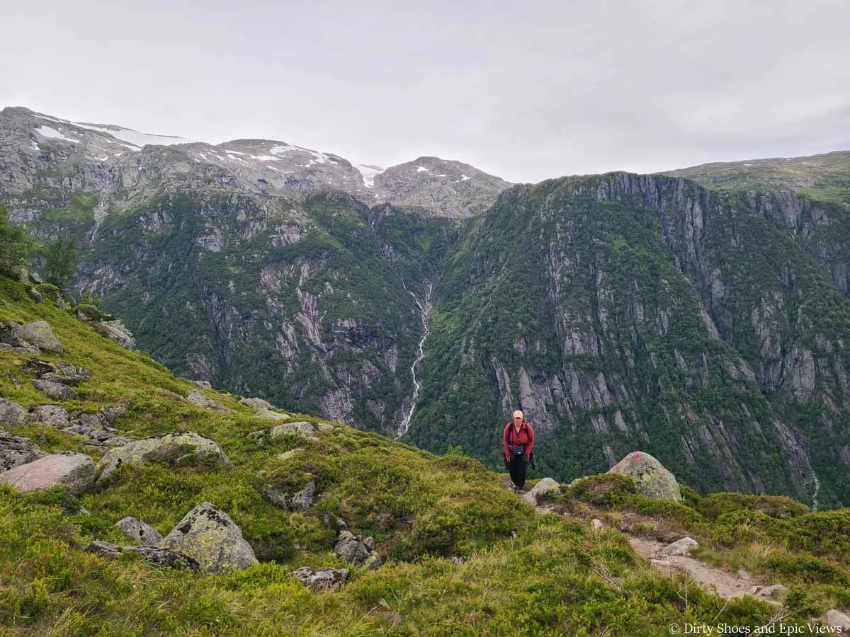 A hiker walks in front of views of snow capped mountains and waterfalls on the Reinanuten trail in Norway