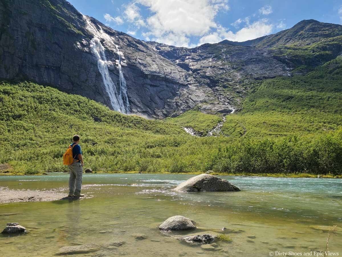 A hiker stands along the shore of a blue stream in front of a waterfall cascading down a rock face near the Briksdal Glacier in Norway