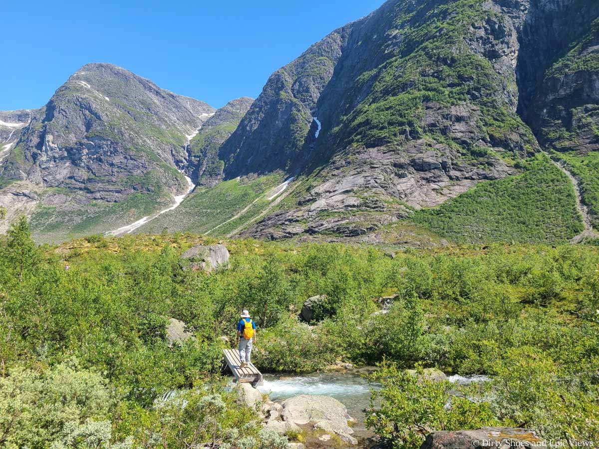 A hiker crosses a footbridge over a blue stream with mountain views on the Austerdalsbreen hike in Norway