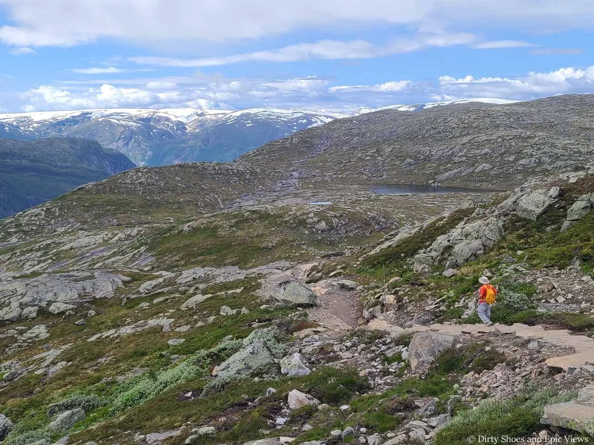 A hiker walks a narrow dirt path through a rocky meadow with mountain views on the Trolltunga trail