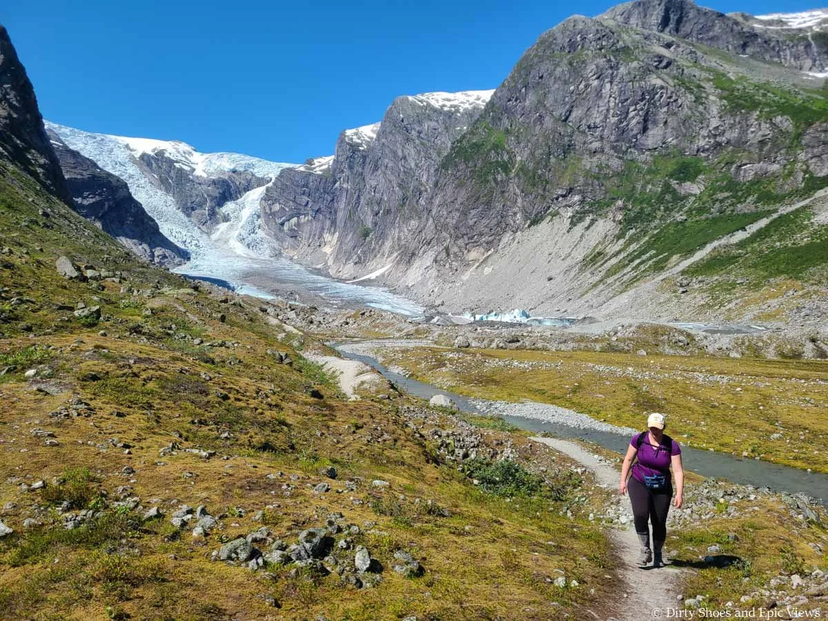 A hiker walks a dirt path along a small stream away from a glacier on the Austerdalsbreen hike