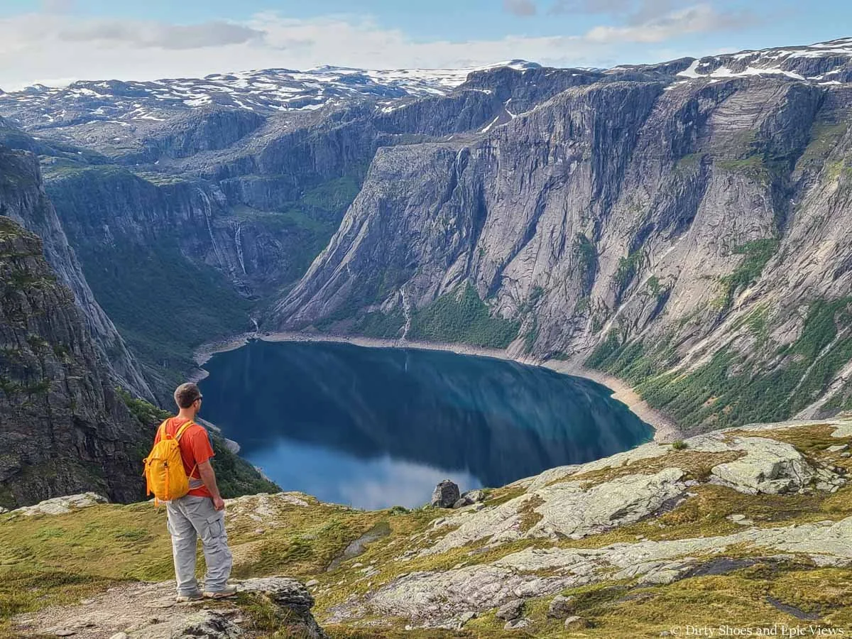A hiker stands on a rocky viewpoint overlooking a deep blue lake surrounded by steep cliffs on the Trolltunga hike