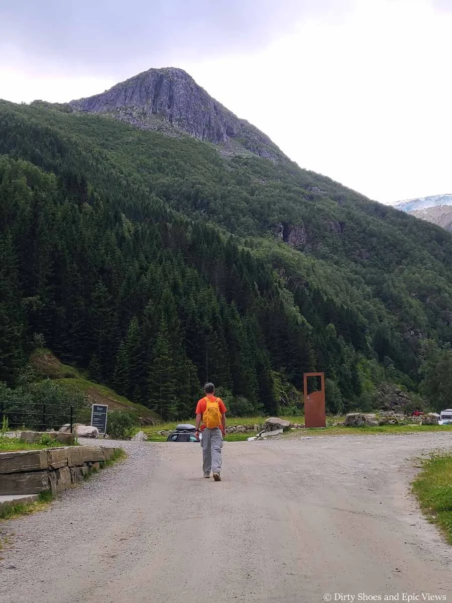 A hiker walks a gravel road beneath tall rocky cliffs in the Reinanuten parking lot in Norway