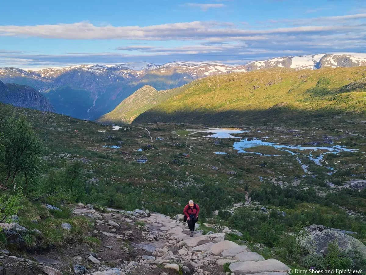 A hiker ascends a stone staircase as morning sun lights up snowcapped mountains in the background with a valley spotted with lakes below