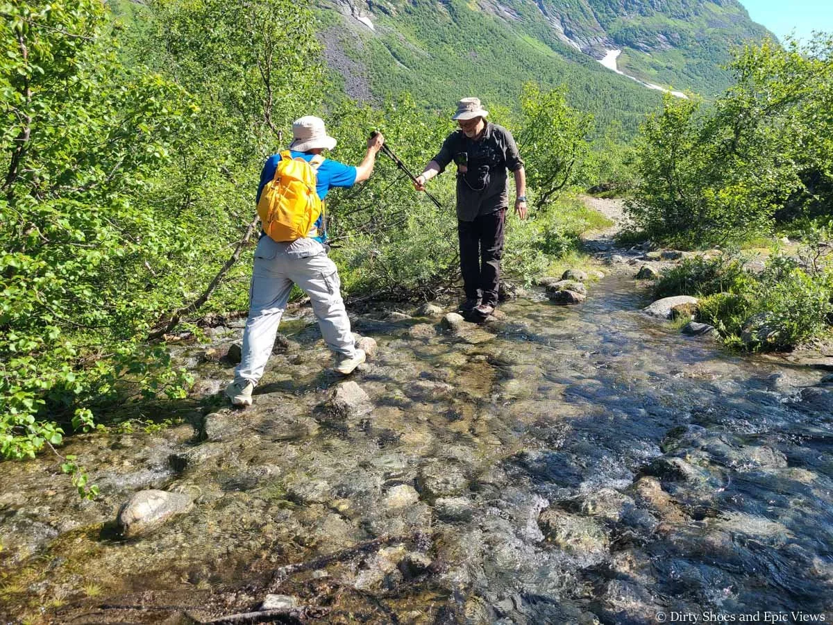 A hiker crosses a stream with the help of a fellow hiker and some trekking poles on the Austerdalsbreen trail