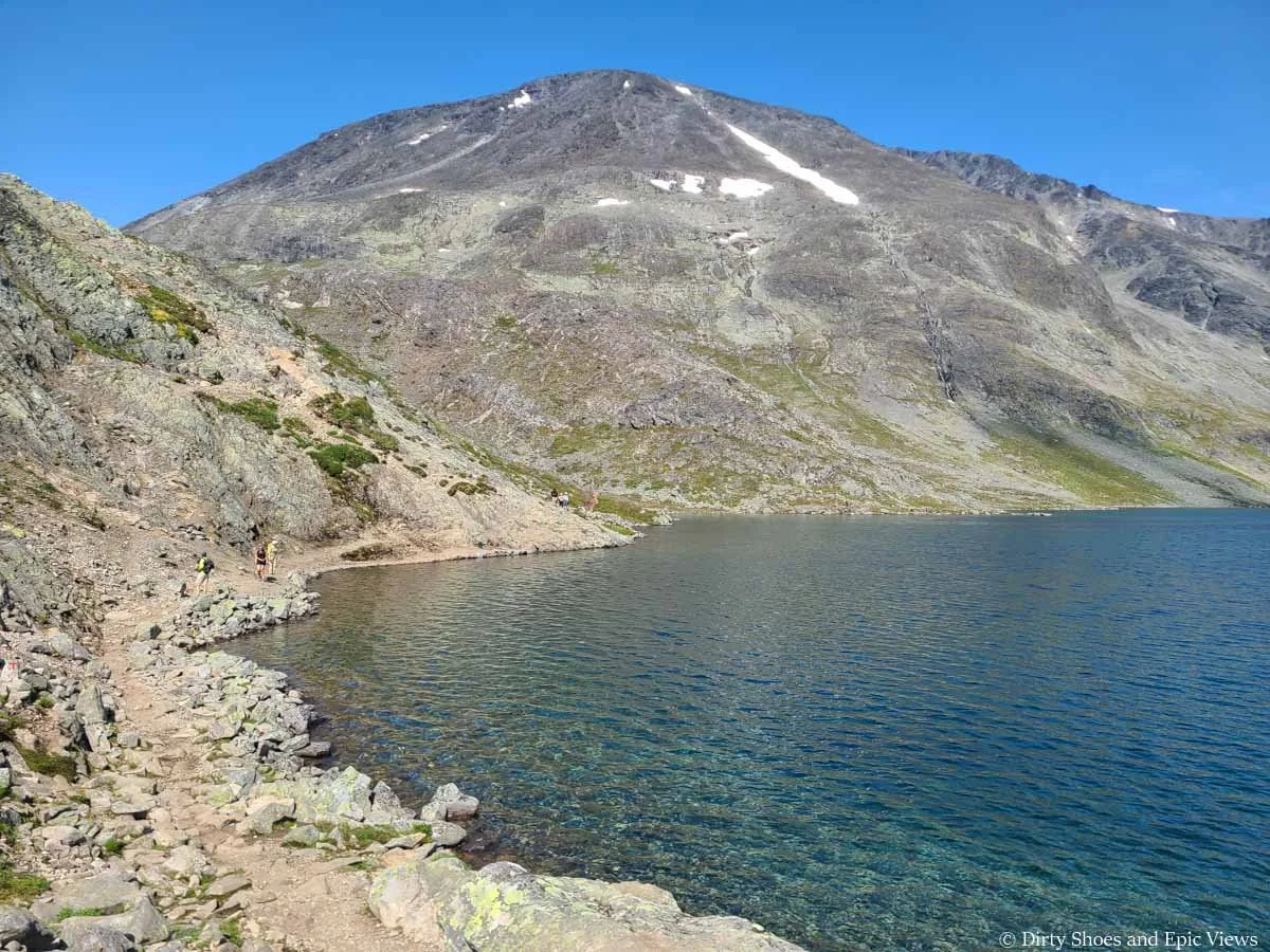 Hikers walk a path around a clear blue lake on the Besseggen Ridge trail in Norway