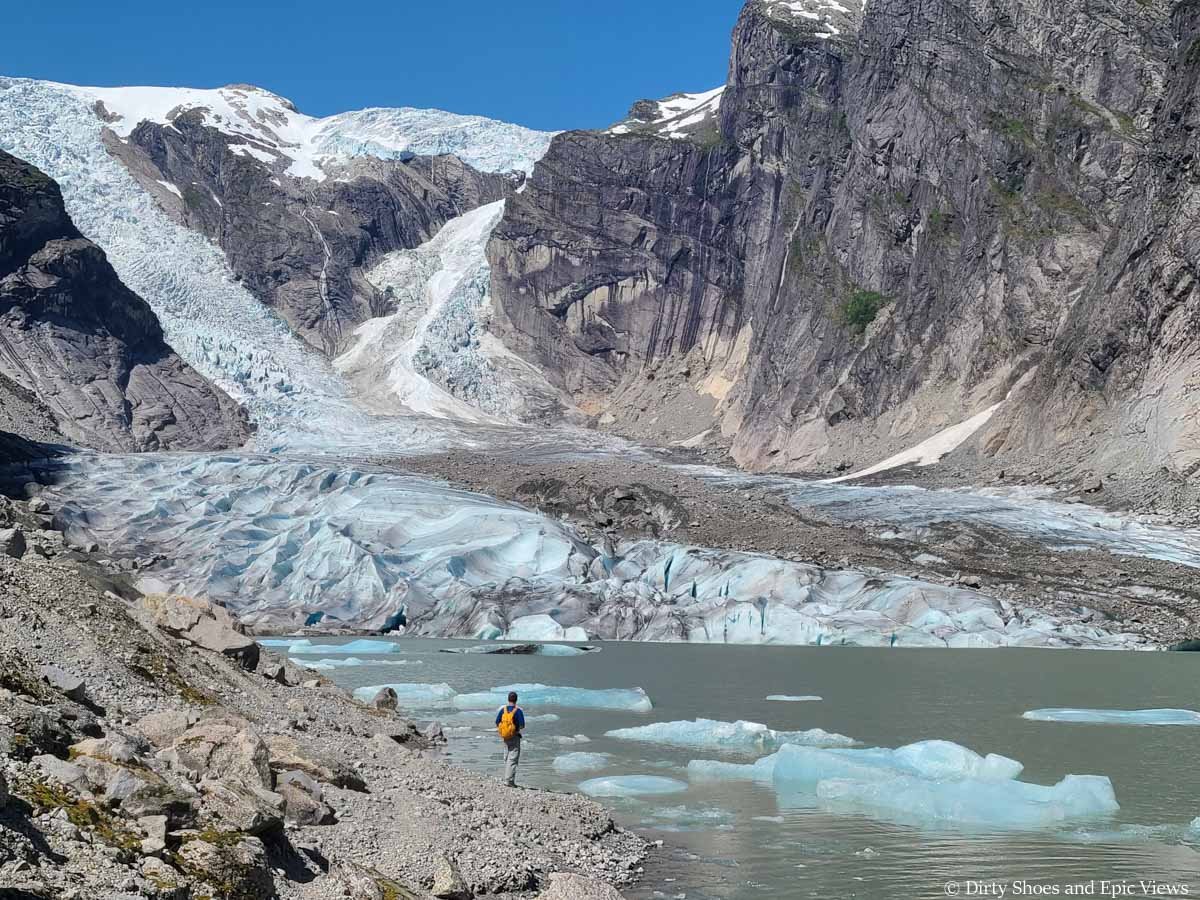 A hiker stands at the shore of a glacial lake looking up at a cascading glacier on the Austerdalsbreen hike in Norway