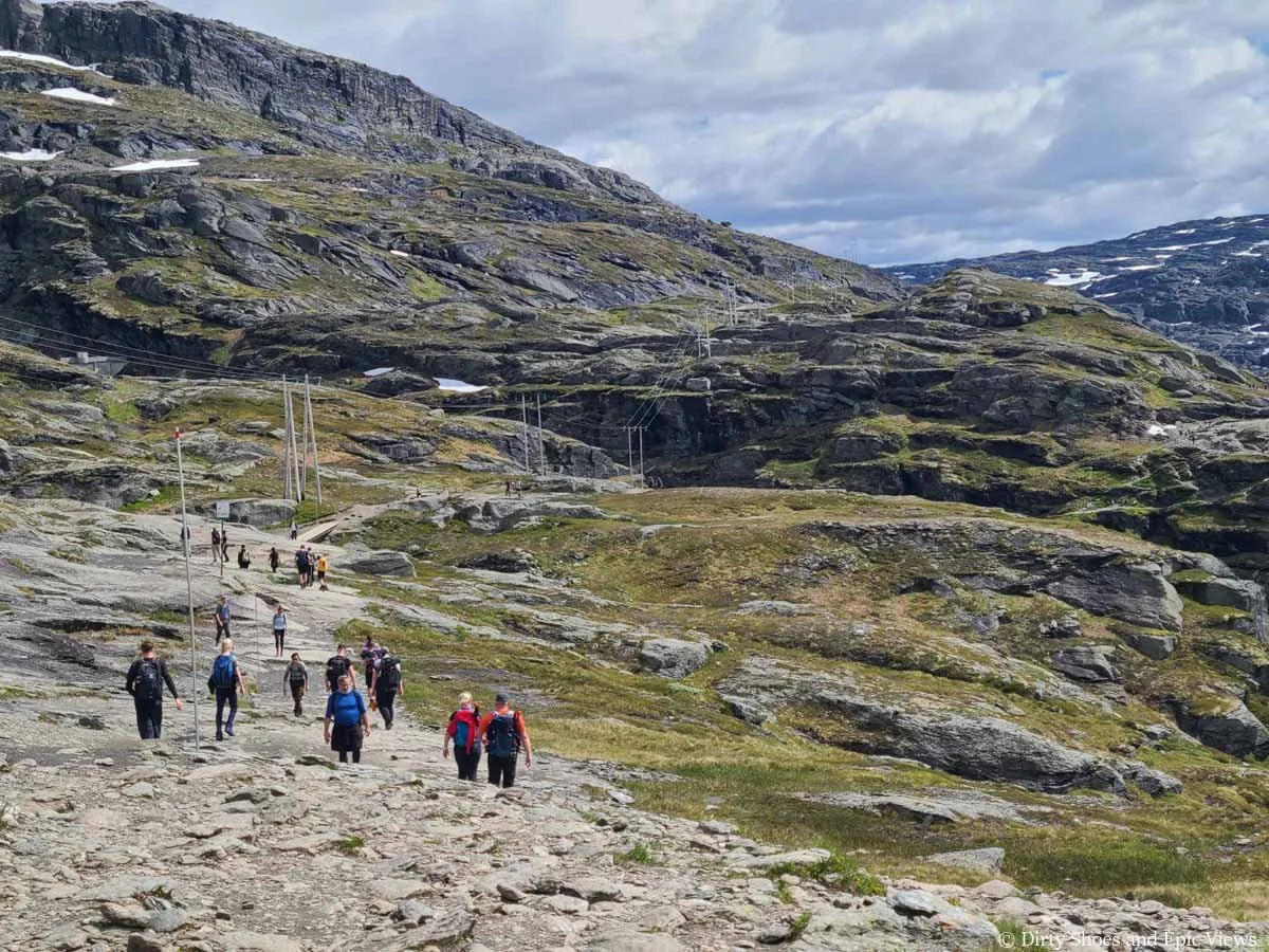 A crowd of hikers walk along a wide rock path through a grassy meadow with mountain views on the Trolltunga hike