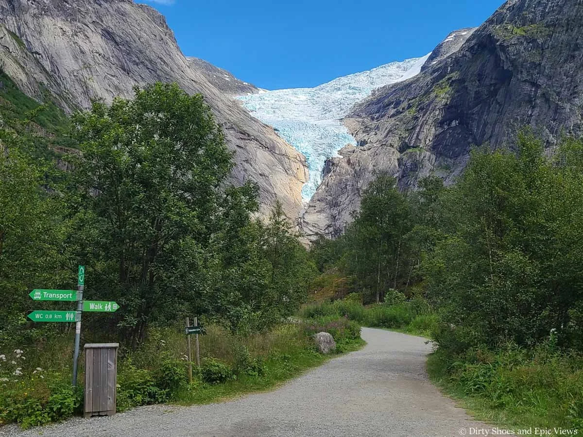 A signpost marks the walking trail as a paved path leads towards a glacier view on the Briksdalsbreen hike in Norway