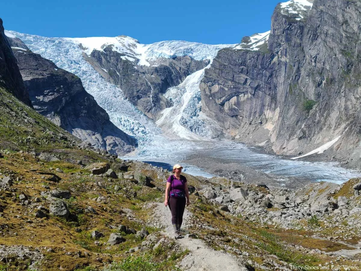 A hiker walks a narrow path through a meadow away from a massive glacier on the Austerdalsbreen hike in Norway