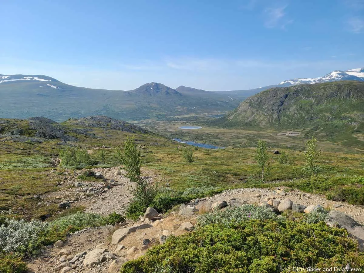 A dirt path weaves through a grassy meadow with distant mountain views on the Besseggen Ridge trail in Norway
