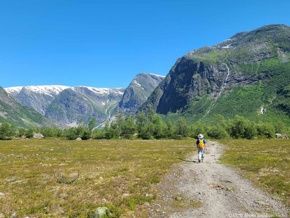 A hiker walks along a wide flat path through a valley surrounded by mountains on the Austerdalsbreen hike in Norway