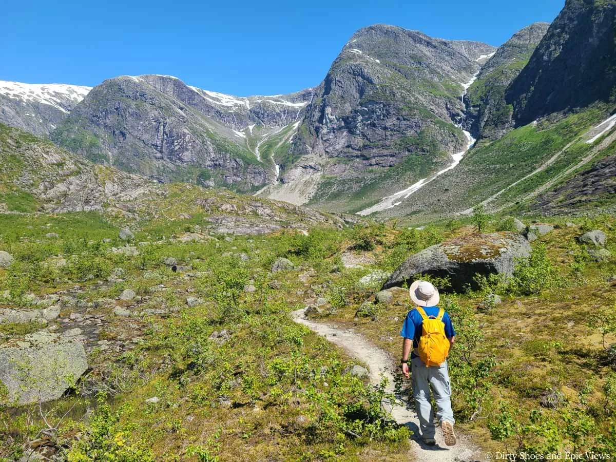 A hiker walks a narrow dirt path through a grassy meadow towards mountain views on the Austerdalsbreen trail in Norway