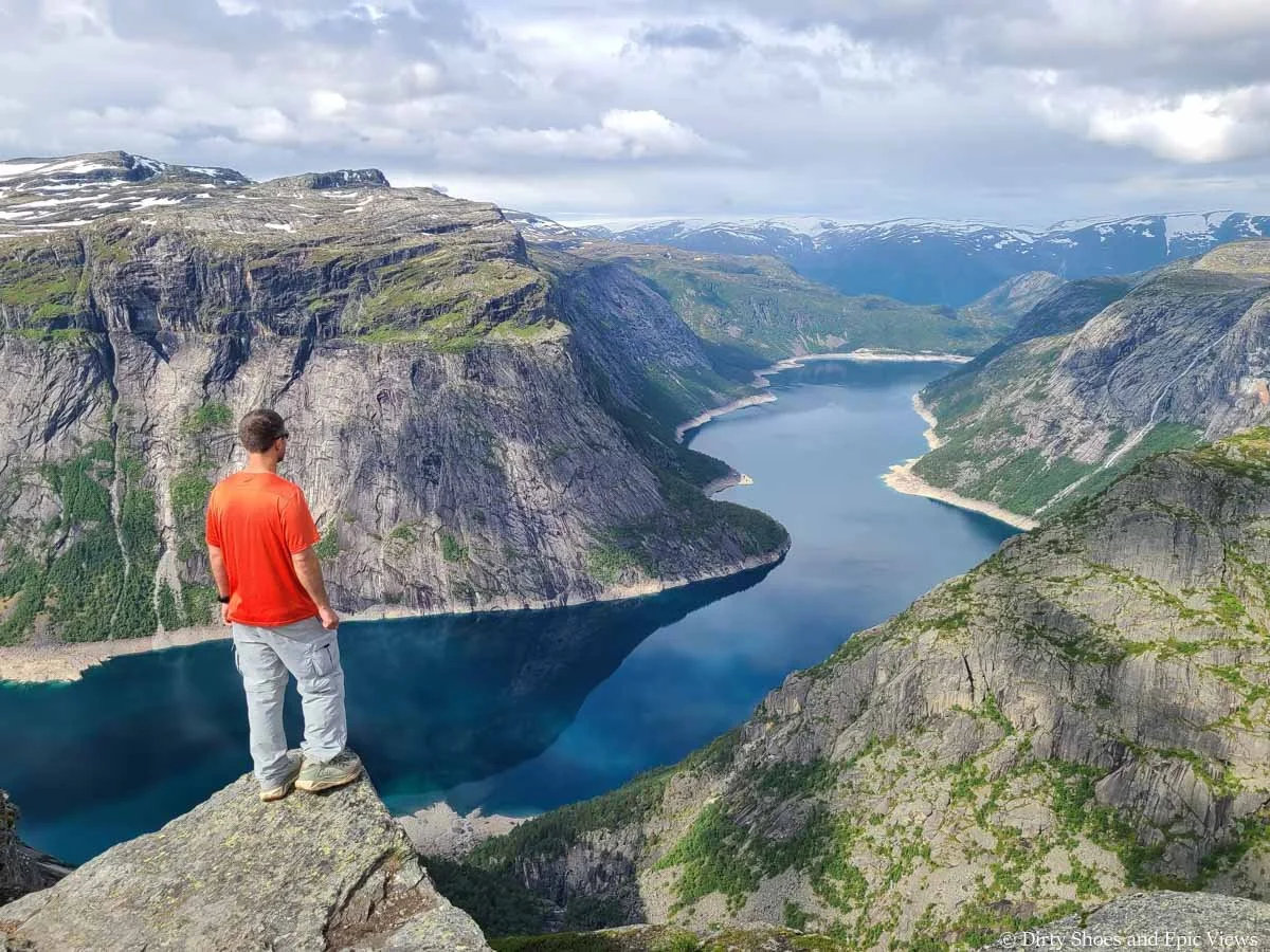 A hiker stands on a rocky overlook above a blue lake surrounded by steep rocky cliffs on the Trolltunga hike