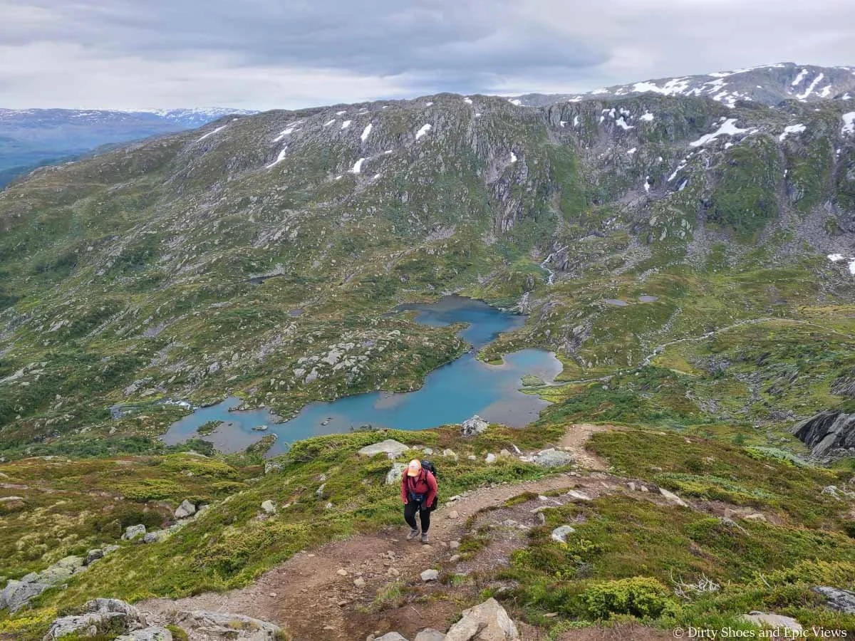 A hiker ascends a steep dirt trail above a basin with blue lakes and waterfalls along the Reinanuten hike in Norway