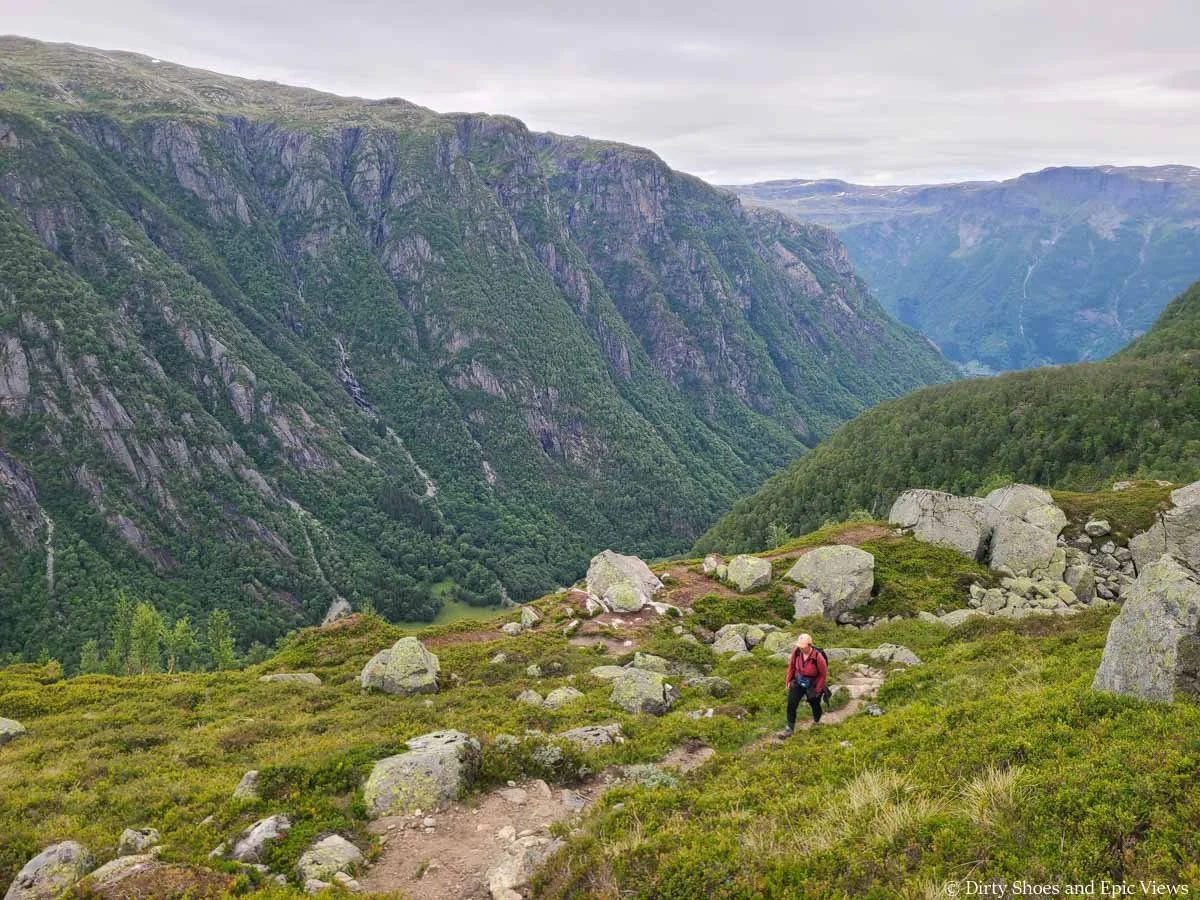 A hiker walks along a dirt path through a grassy meadow with mountain views on the Reinanuten trail in Norway