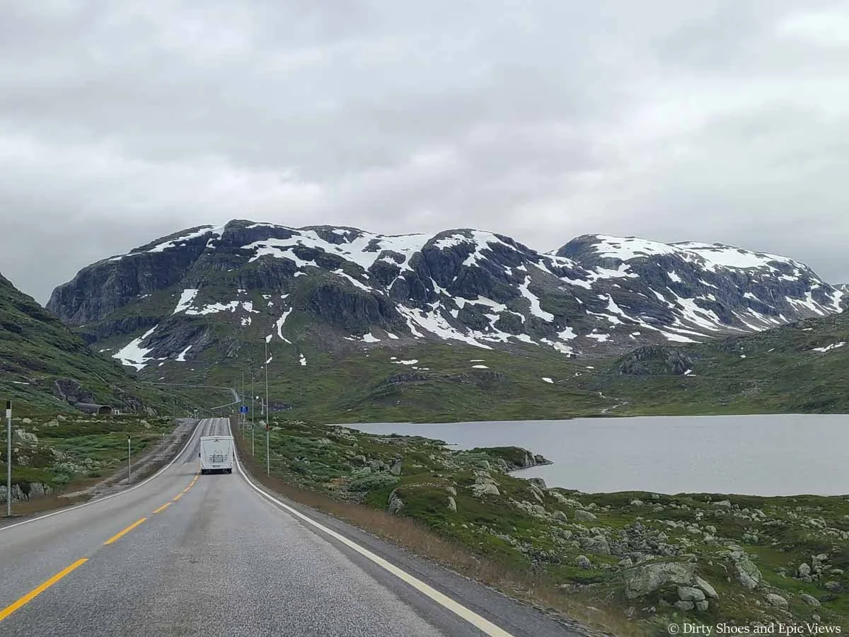 A campervan drives a road towards snow covered mountains along lake on the drive between Oslo and Odda