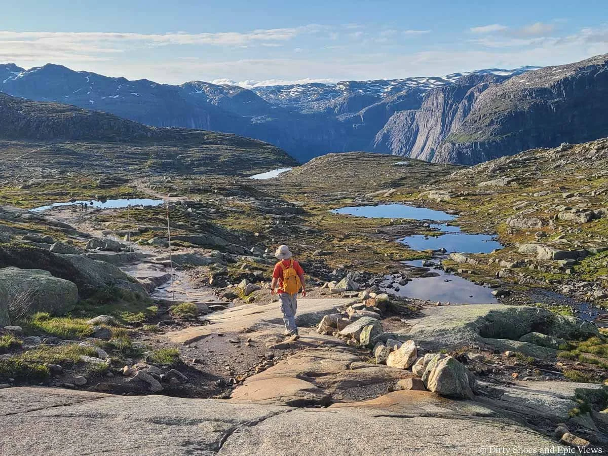 A hiker descends a rocky trail towards a meadow filled with lakes and backdropped by mountains on the Trolltunga trail