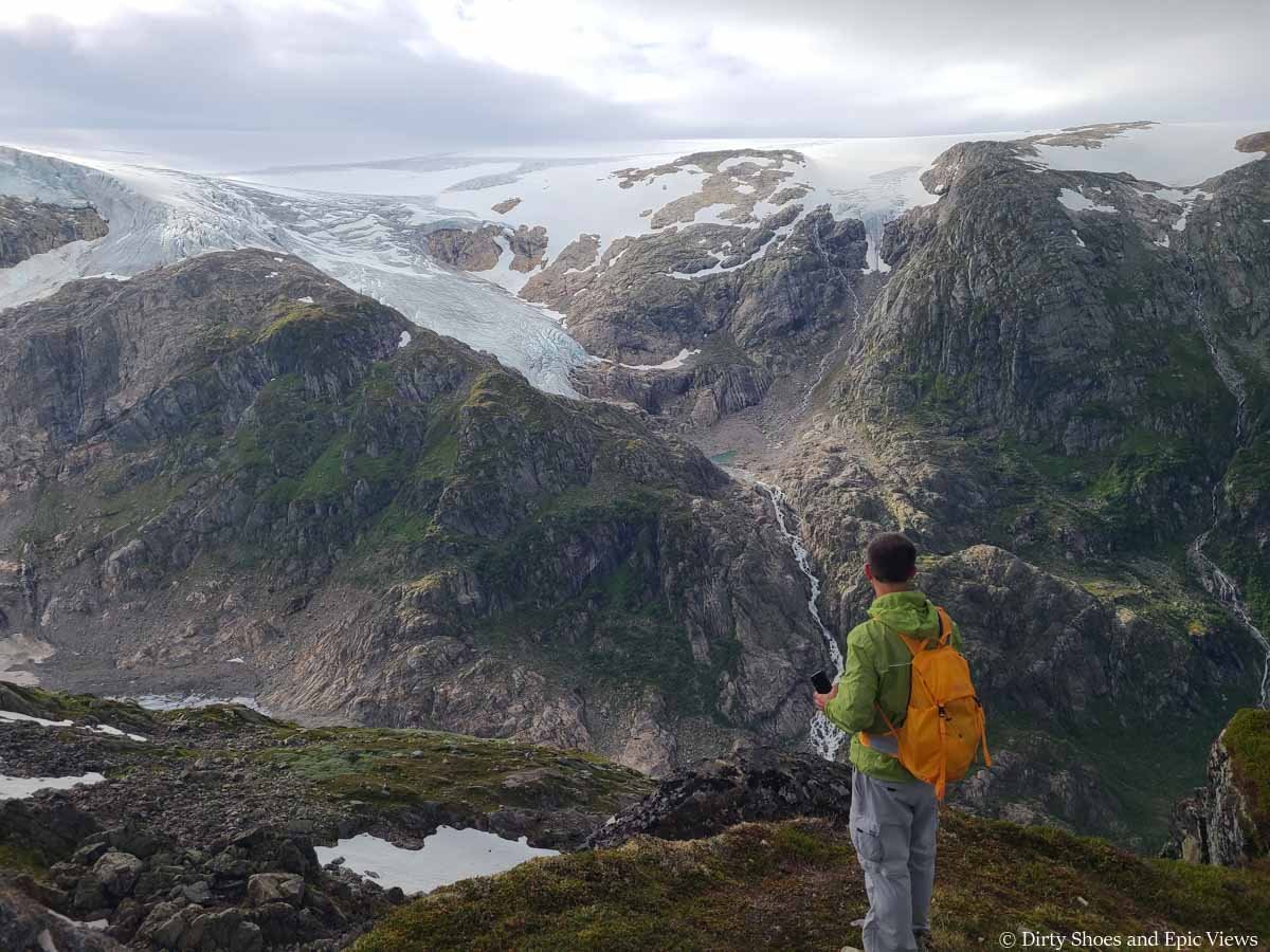 A hiker stands in front of views of snow covered cliffs and waterfalls along the Reinanuten hike in Norway