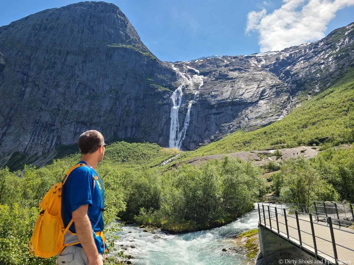 A hiker looks out over a blue river to a waterfall streaming down a rock face on the Briksdal Glacier trail