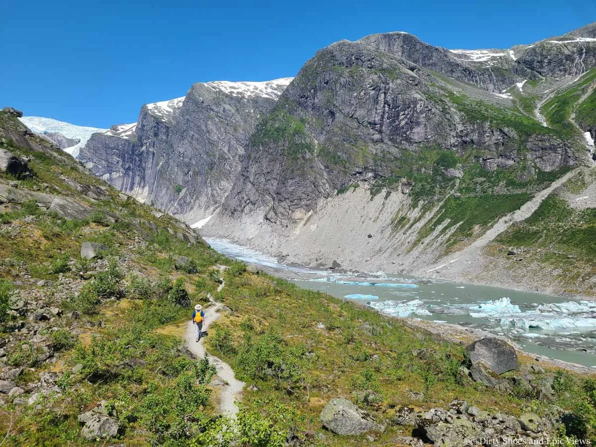 A hiker walks a narrow dirt path along a grassy slope above a glacier lake on the Austerdalsbreen trail in Norway