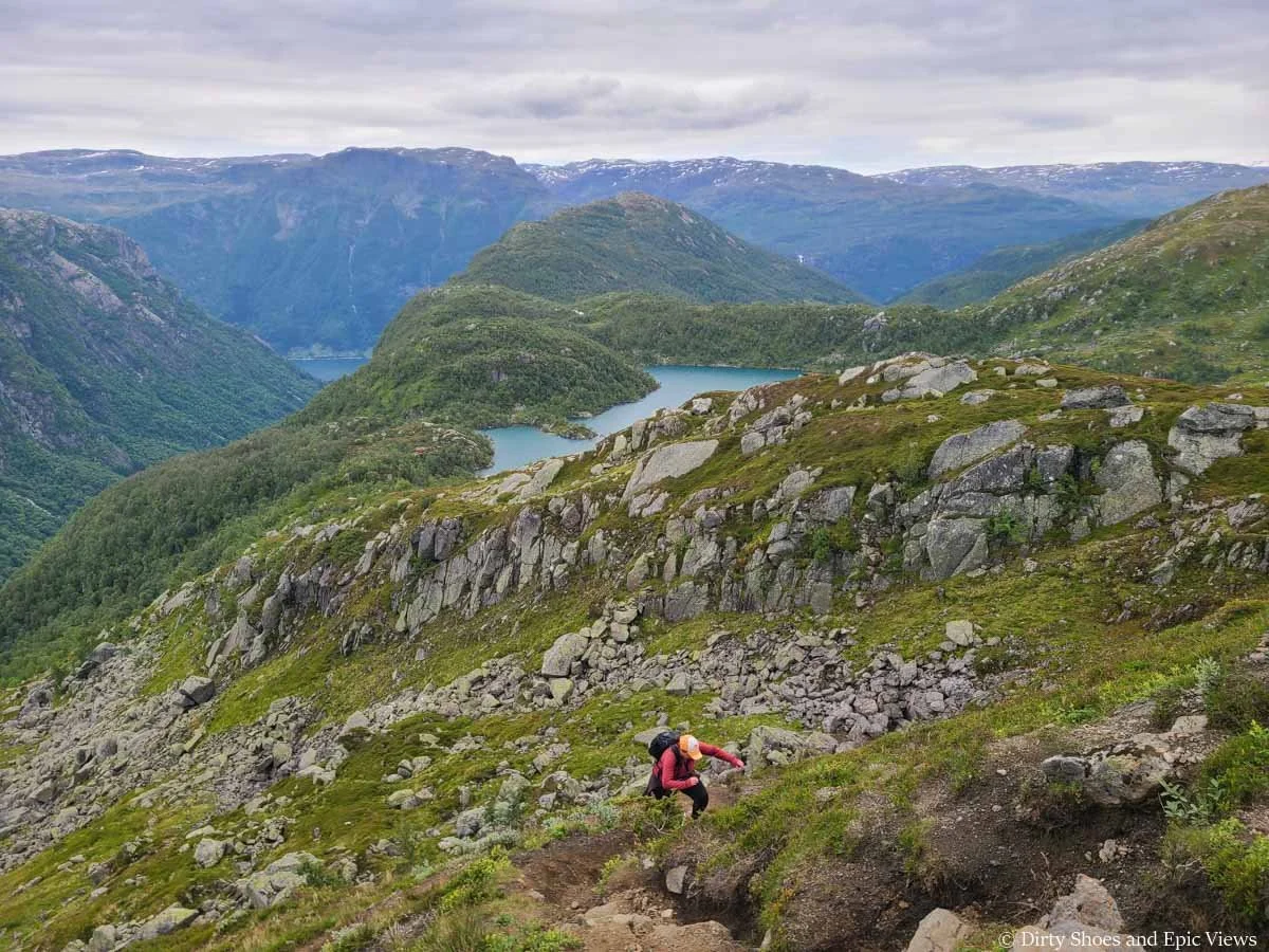 A hiker ascends a steep path up a meadow with views of mountains and lakes on the Reinanuten trail in Norway