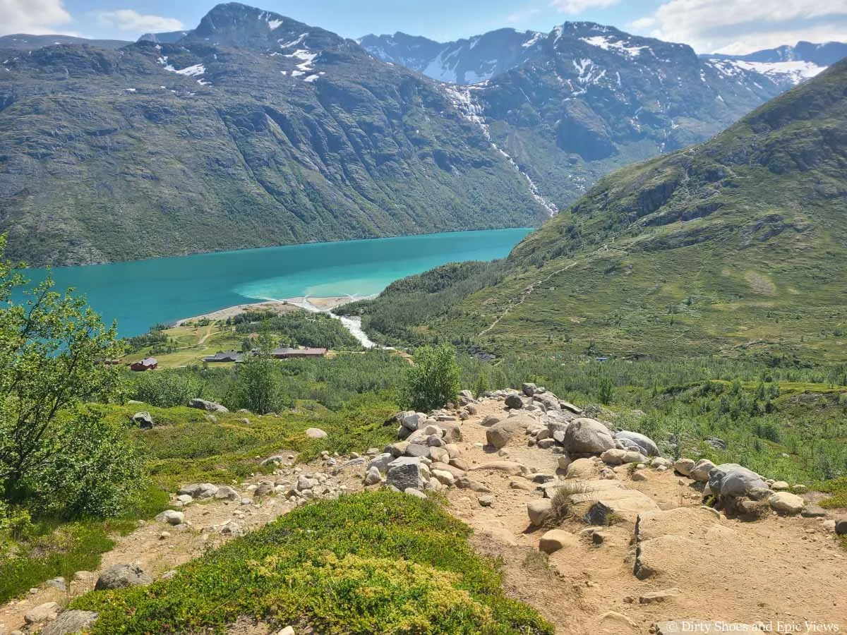 A rocky trail descends steeply towards buildings at the edge of a blue lake on the Besseggen Ridge trail