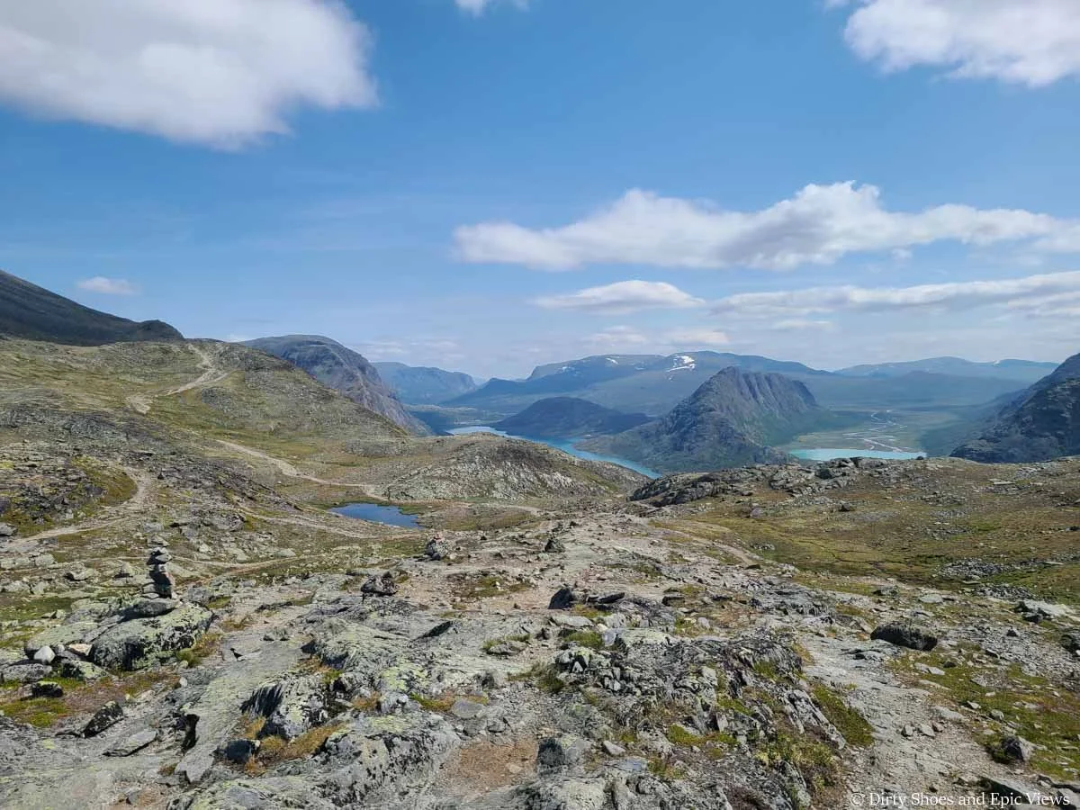 A path descends gently down a grassy and rocky slope towards mountain and lake views on Besseggen Ridge